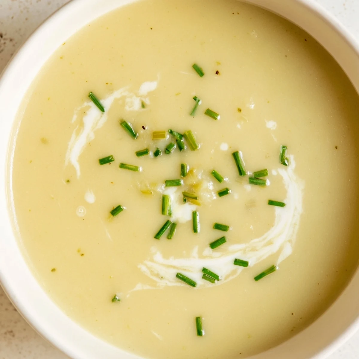 Creamy potato leek soup in a white bowl topped with chopped chives, served beside a linen napkin and a wooden spoon.