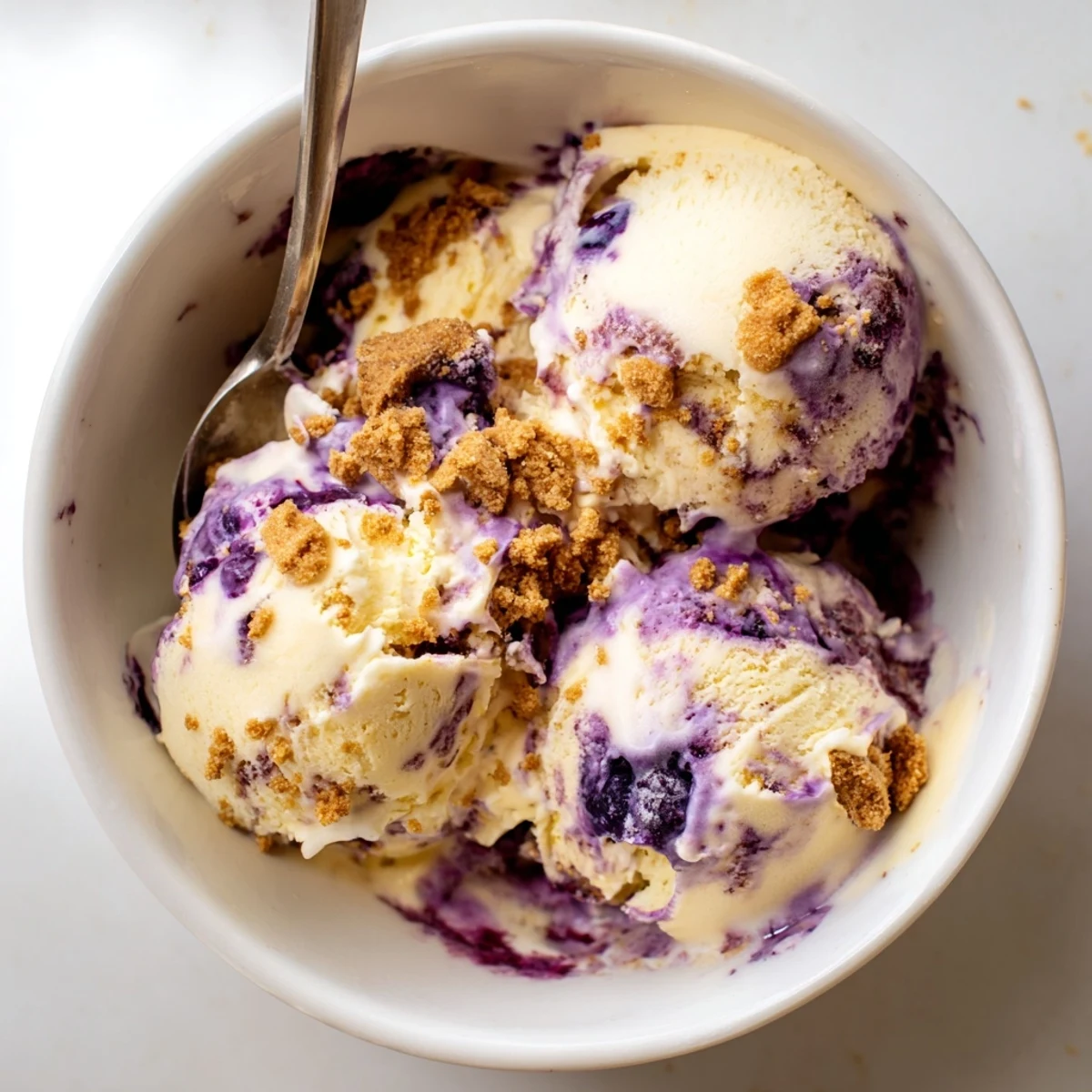 A scoop of Lemon Blueberry Cheesecake Ice Cream in a clear glass bowl, showcasing creamy texture and blueberry swirls with graham cracker crumbs.