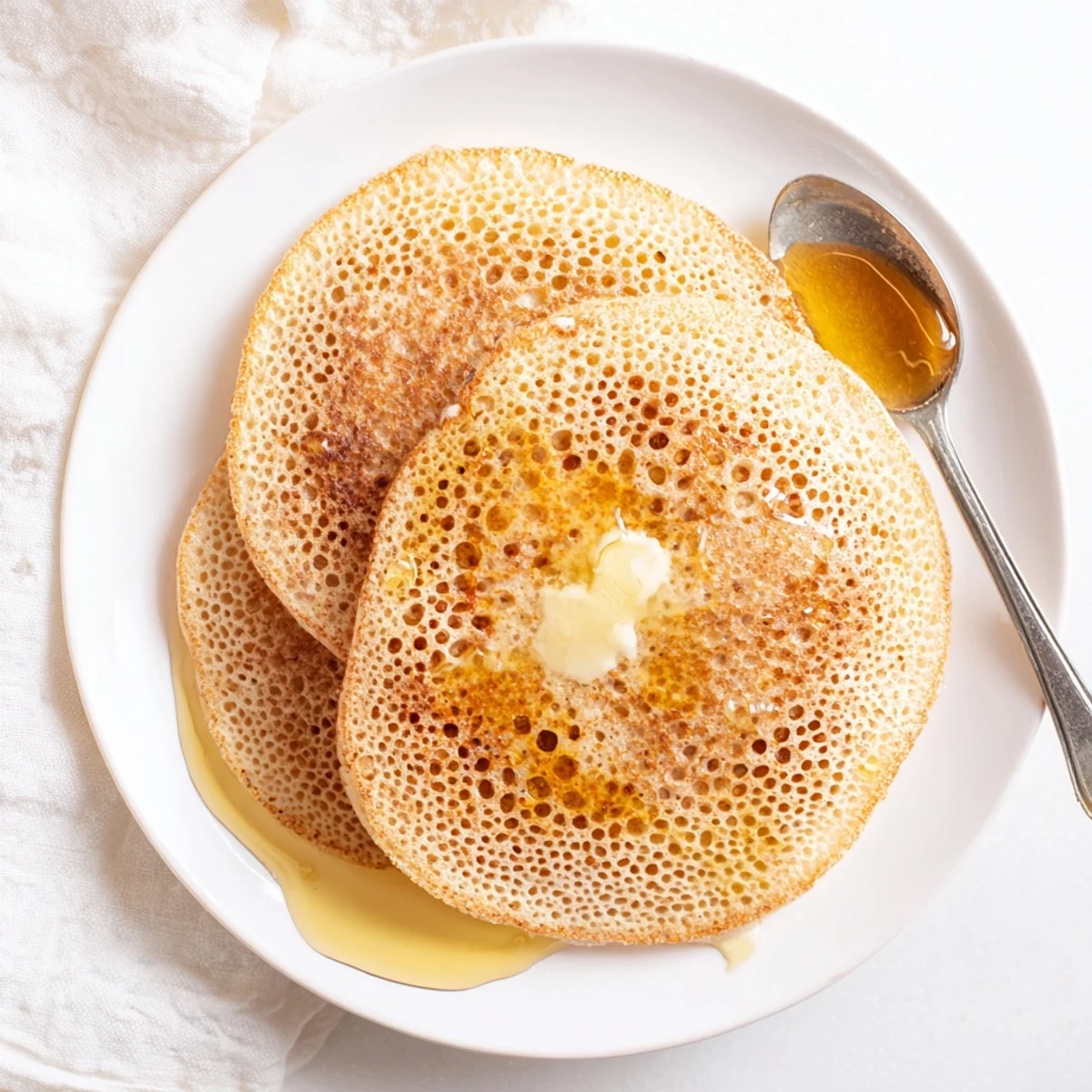 Stack of spongy Baghrir Moroccan pancakes topped with butter and honey on a white plate