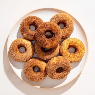 Twelve soft, fluffy Baked Cinnamon Sugar Donuts You Can't Resist arranged on a cooling rack with a jar of cinnamon sugar nearby.