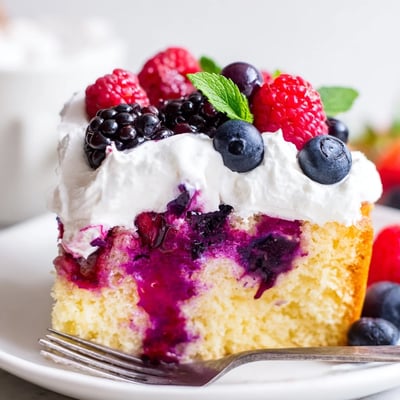 Moist Summer Berry Poke Cake drizzled with vibrant berry sauce on a white plate